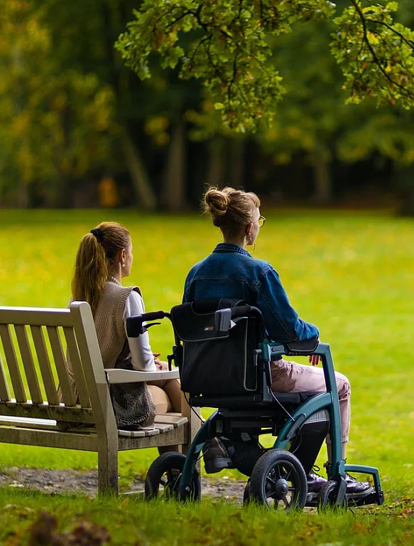 Zwei Frauen sind von hinten in einem Park fotografiert. Die eine Frau sitzt auf einer Bank, die andere Frau sitzt auf einem Rollator.