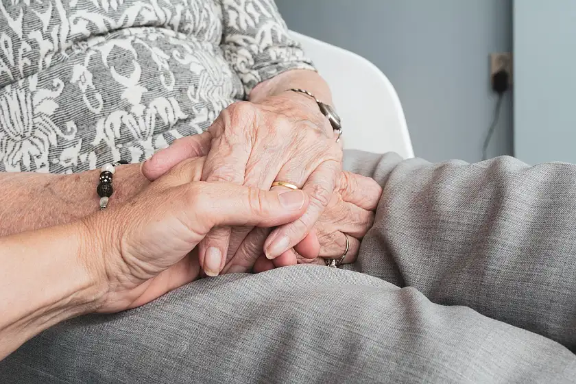 Foto einer älteren Person, die sitzt und im Schoß die Hand einer jüngeren Person hält. Das Foto zeigt keine Gesichter, sondern nur die haltenden Hände.