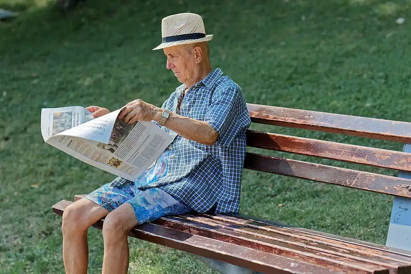 Ein Senior sitzt auf einer Parkbank im Schatten in luftiger Kleidung und mit Sonnenhut. Er liest Zeitung