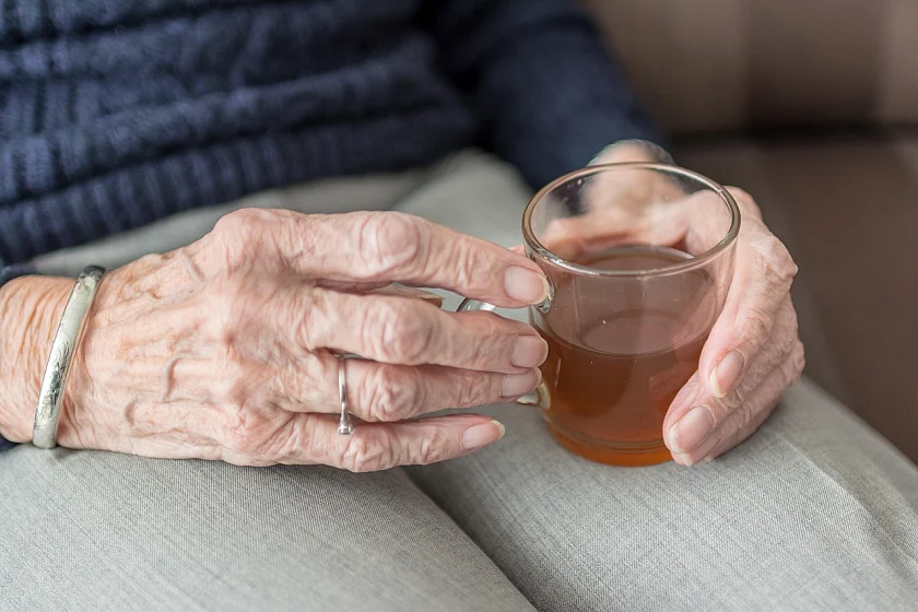 An elderly woman sits in an armchair and holds a glass of tea in her lap.