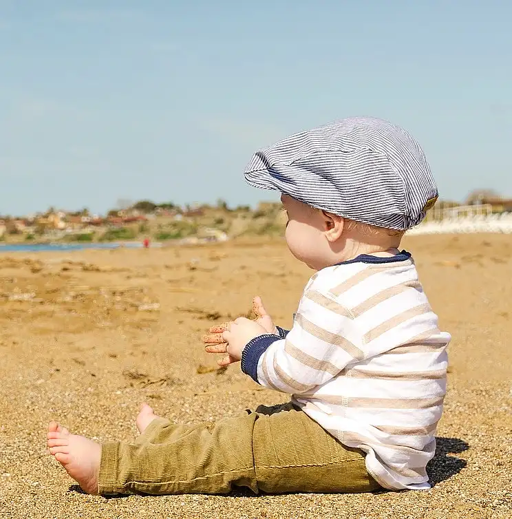 A toddler sits on a beach, dressed in long clothing and wearing a hat.