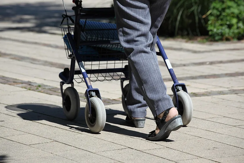 Ein Foto einer Person, die bei sonnigem Wetter mit einem Rollator spaziert. Nur die Beine der Person sind zu sehen.