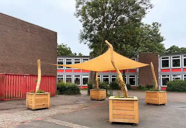 Sonnensegel mit Holzpfählen und bepflanzten Kübeln auf einem Schulhof