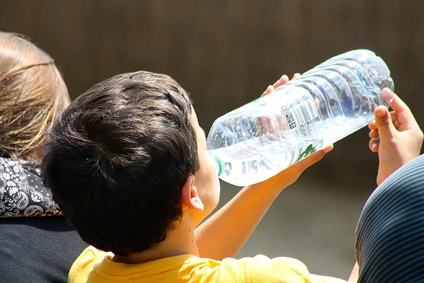 Ein Junge ist von hinten zu sehen, wie er bei Sonneneinstrahlung Wasser aus einer Flasche trinkt.